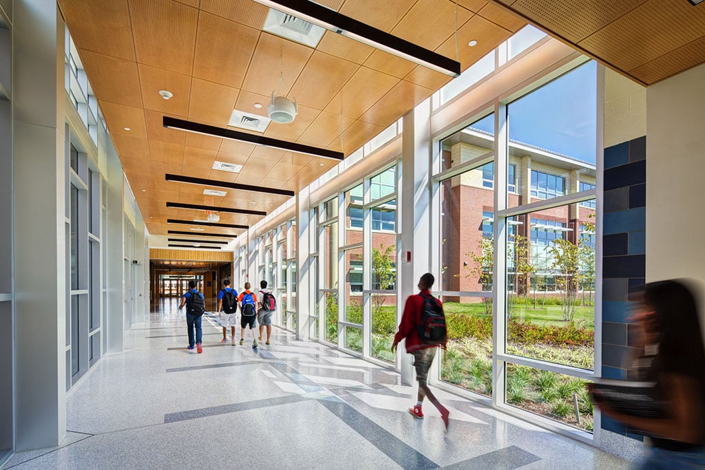 A bright high school hallway showing a green courtyard on a sunny day through floor to ceiling windows as students walk through. 